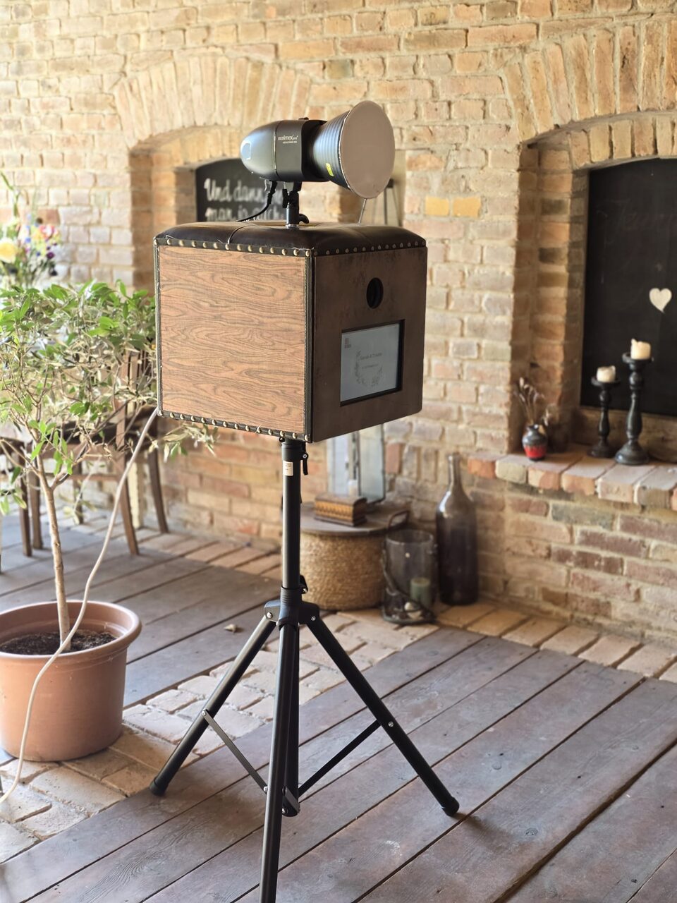 Die Fotobox aus Holz und Leder auf einem Stativ, darüber ein Blitzkopf, dahinter eine Backsteinwand