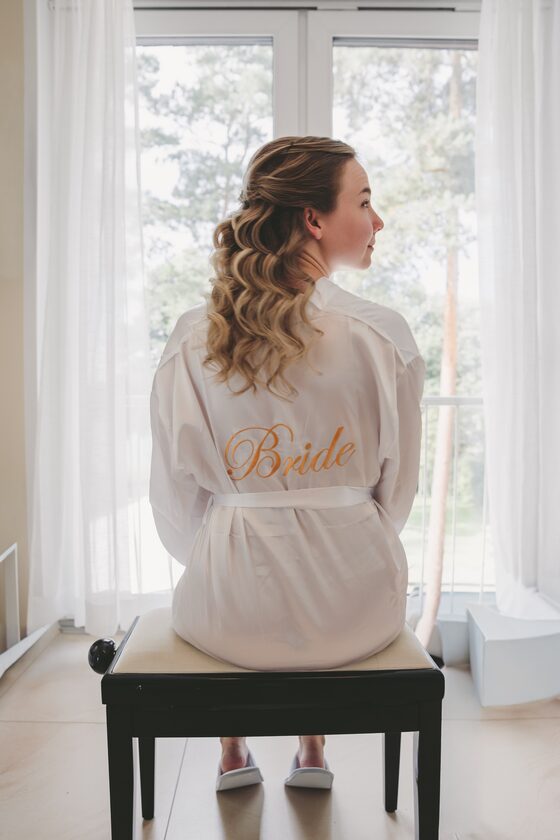 Braut im Morgenmantel mit dem aufgestickten Schriftzug „Bride“ sitzt am Fenster und blickt hinaus